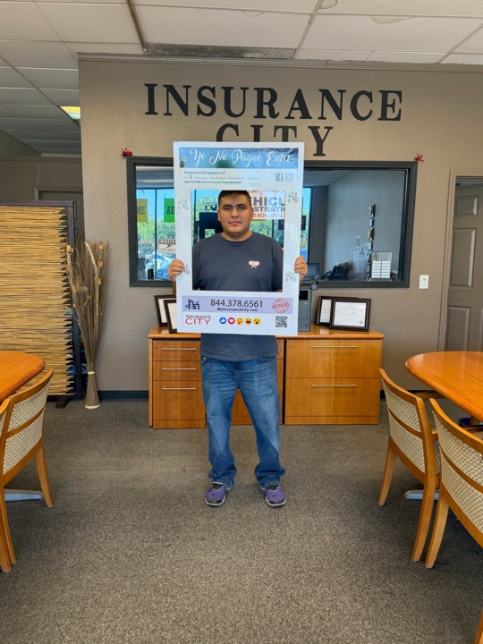Insurance City Stockton customer holding Yo No Pague Entre frame in front of office signage — no broker fee paid